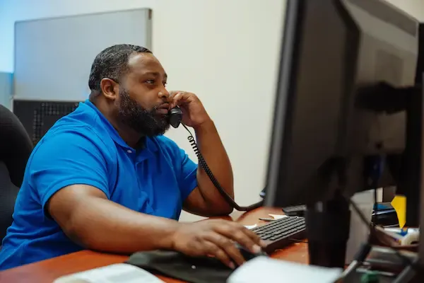A bearded man in a blue polo shirt sits at an office desk, talking on a landline phone while looking at a computer monitor and using a mouse.